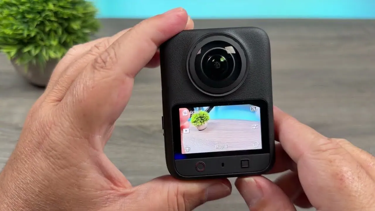 Two hands holding the DJI Osmo 360 II with its lens and touchscreen facing forward, the screen showing an 8K 30 frames per second preview of a potted plant on a desk.
