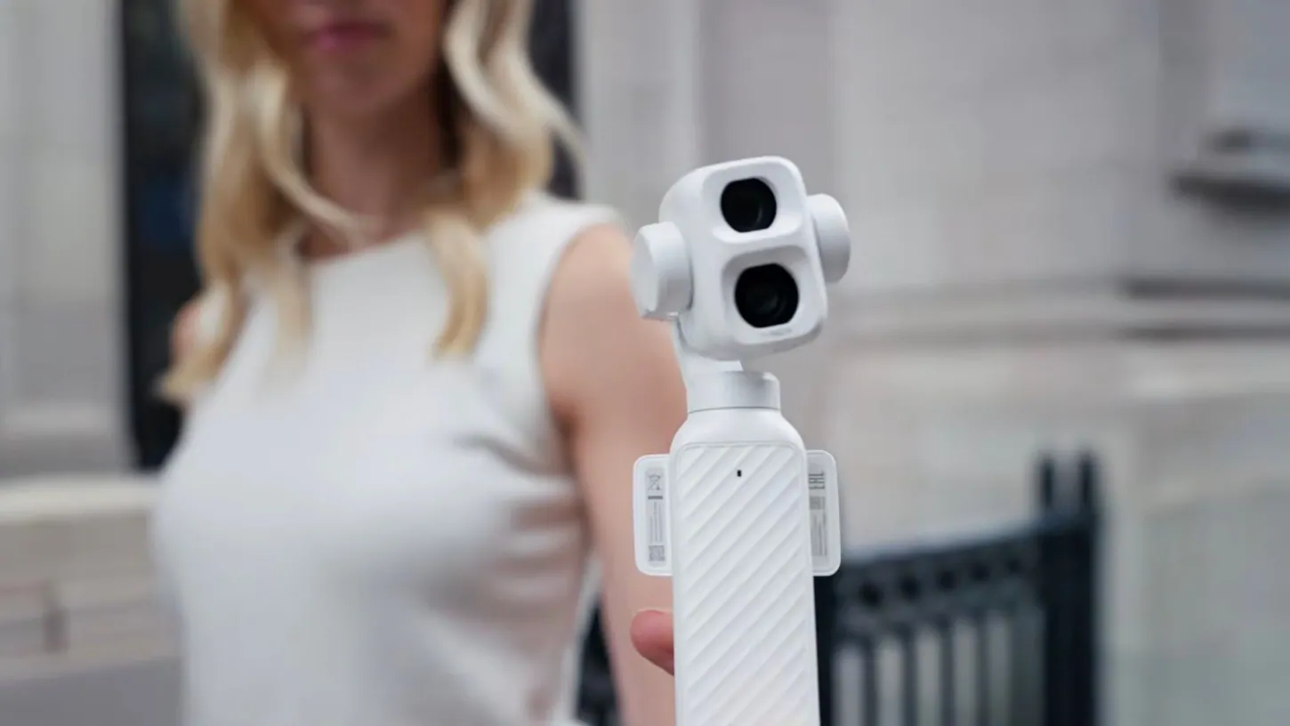 White DJI Osmo Pocket 4P with its two stacked camera lenses held out toward the viewer by a woman in a white top, out of focus on a city street