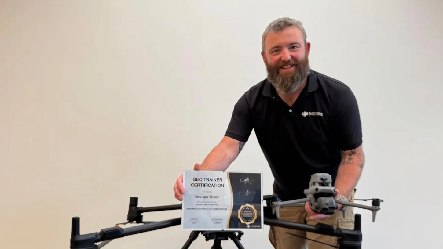 Bearded man in a black DJI Enterprise polo holding a GEO Trainer Certification and a DJI Matrice 4E, with a DJI Matrice 400 on a wooden table in front of him