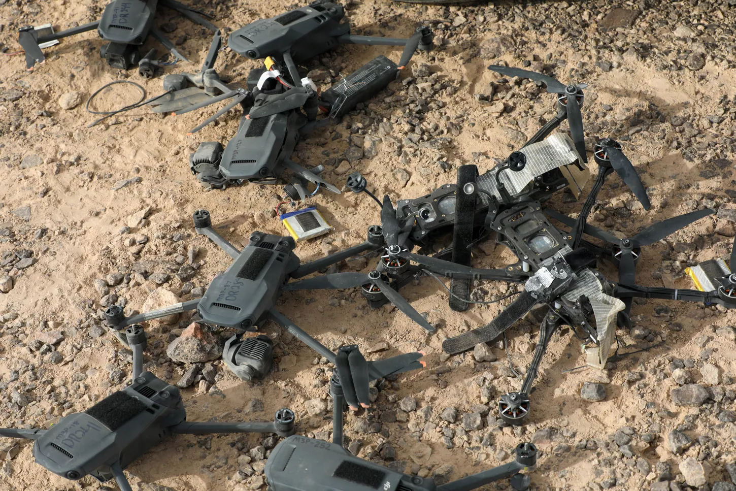 Several gray folding quadcopters and a black FPV drone frame lie damaged on rocky desert sand.