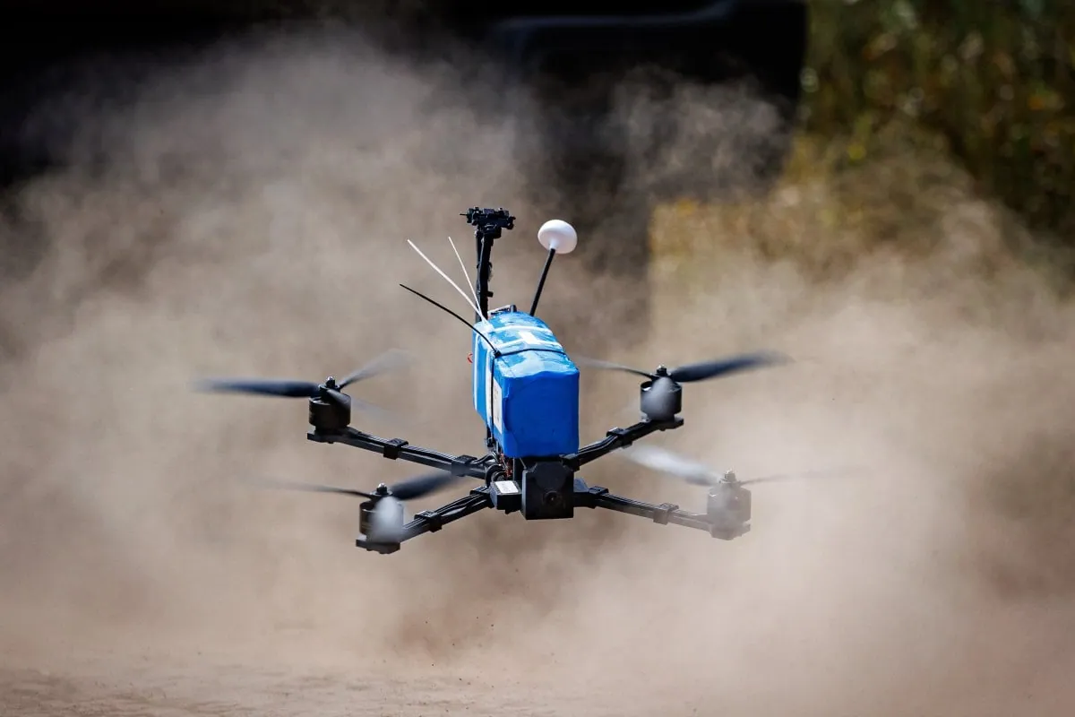 Blue-bodied Lupynis-10 FPV quadcopter lifting off a dirt track in a cloud of dust, with a white GPS puck and antennas on top