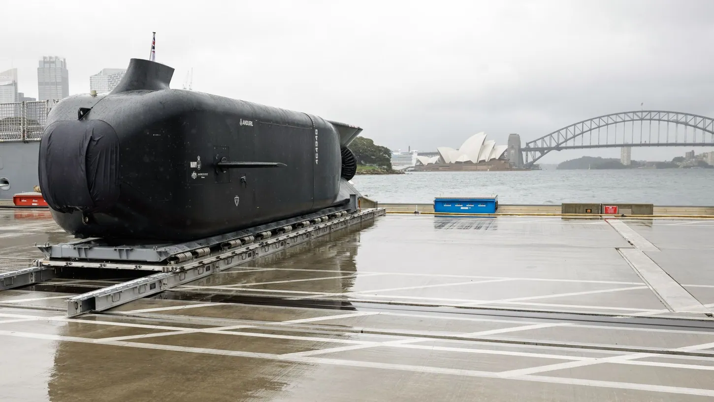 Black Anduril Ghost Shark extra-large autonomous underwater vehicle on a cradle at a Sydney wharf, with the Opera House and Harbour Bridge behind it