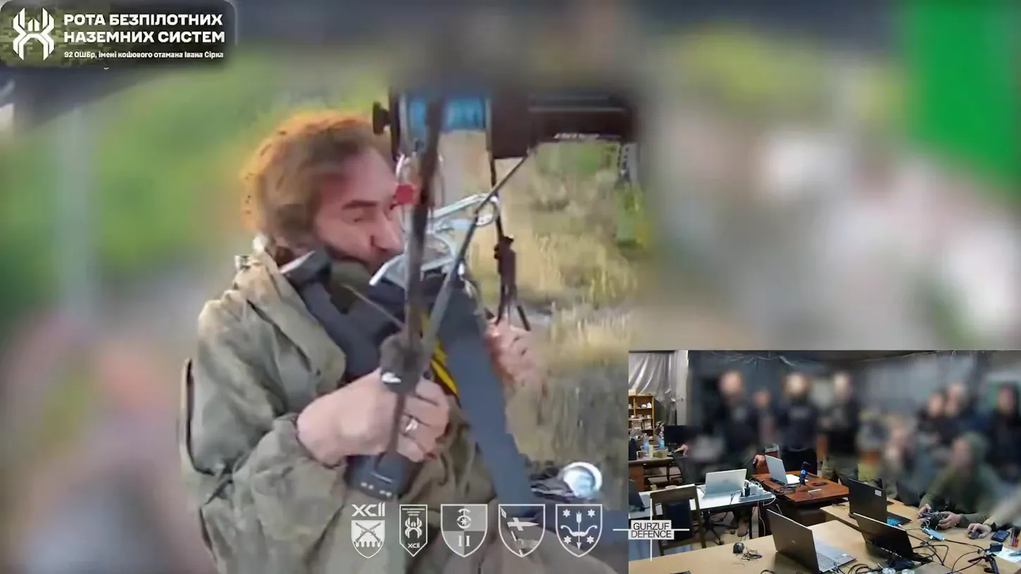 Camera view from the drone looking down at a bearded soldier in a harness gripping the rigging, with an inset of operators at laptops in a command post
