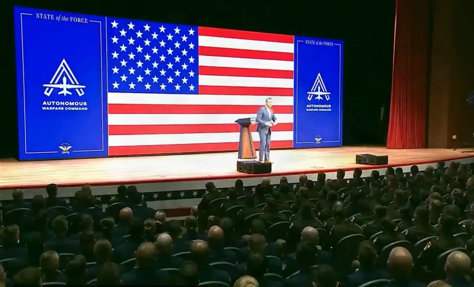 Wide view of a theater stage with a large U.S. flag screen, Pete Hegseth at a lectern, Autonomous Warfare Command logos on blue panels either side, and rows of uniformed troops seated below