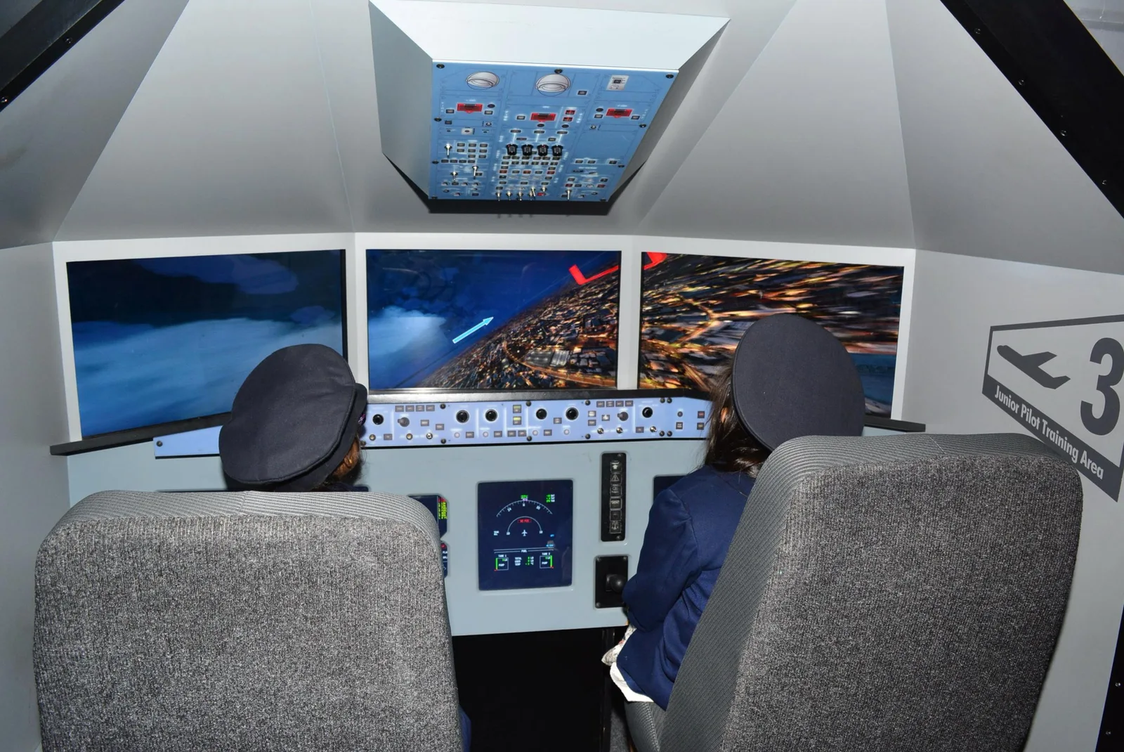 Two children in pilot caps sitting at a junior flight simulator cockpit with multiple display screens