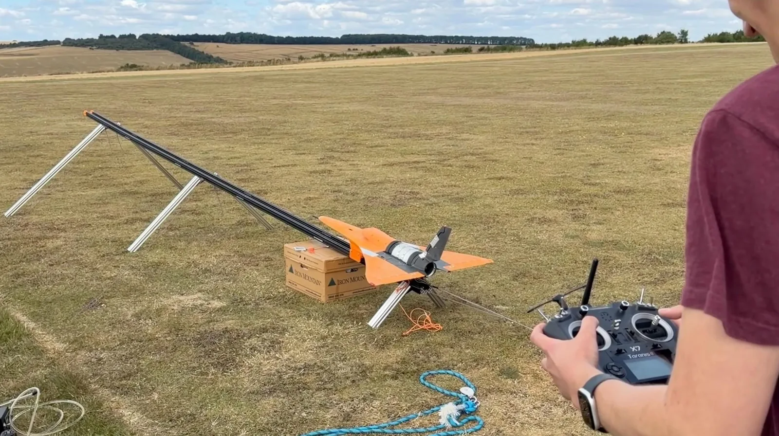 A pilot holding a FrSky Taranis X7 radio transmitter in the foreground, with an orange jet aircraft loaded on a long bungee launch rail on a grass airfield