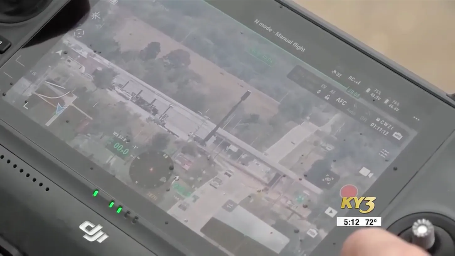 Close-up of a DJI remote controller screen showing an aerial view of a road and buildings, with flight mode, wind speed and two battery readouts on the display