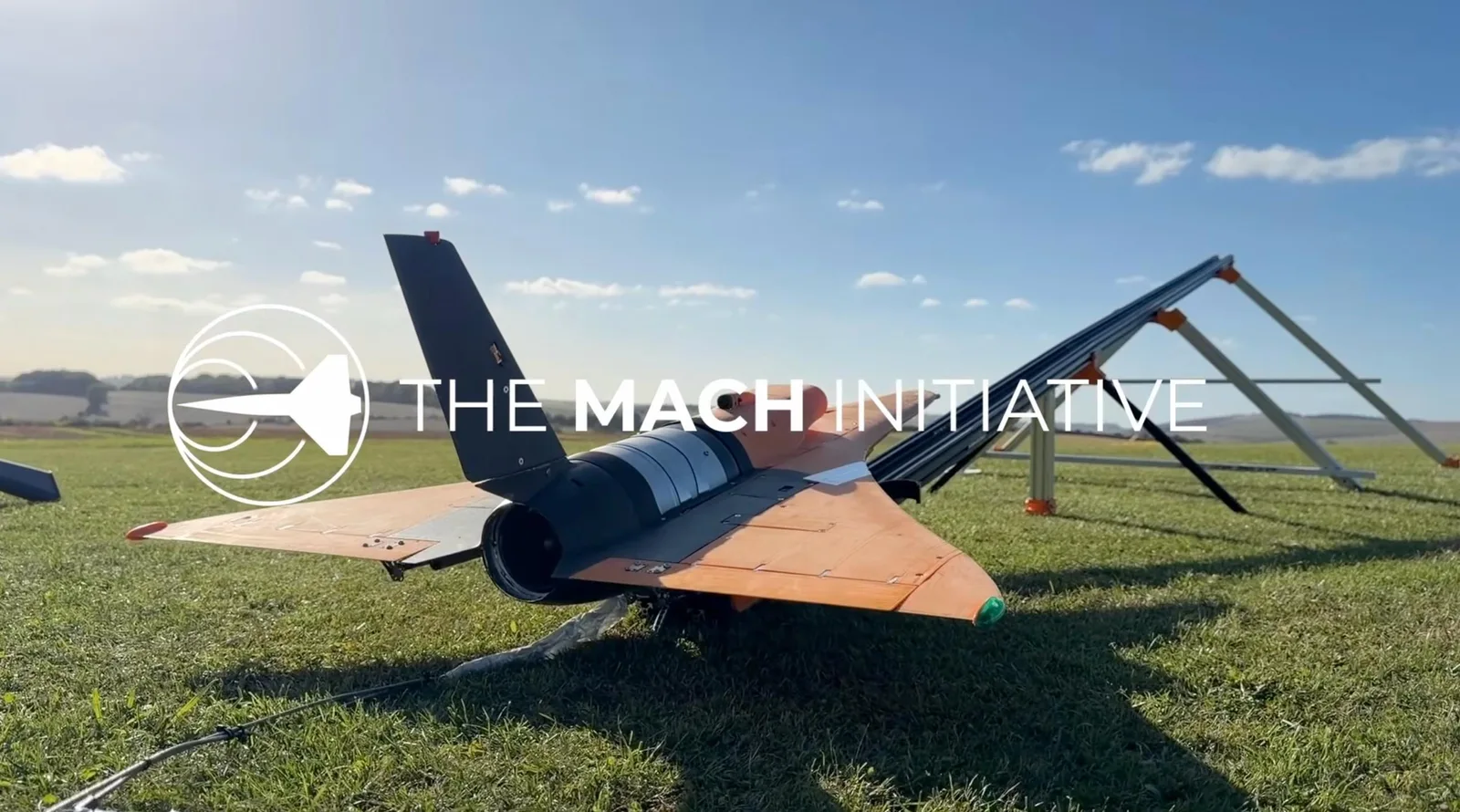 Rear view of The Mach Initiative's orange jet aircraft on grass with its launcher ramp behind it, overlaid with the team's logo and name