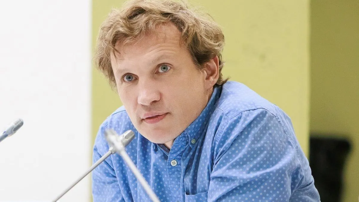 Maksym Savanevskyi in a blue patterned shirt leaning on a conference table between microphones