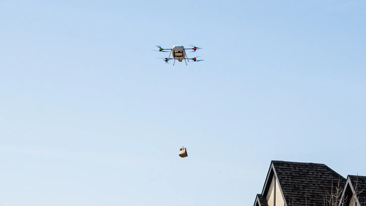 A Manna quadcopter hovers in a blue sky above a house roof while lowering a paper delivery bag on a tether
