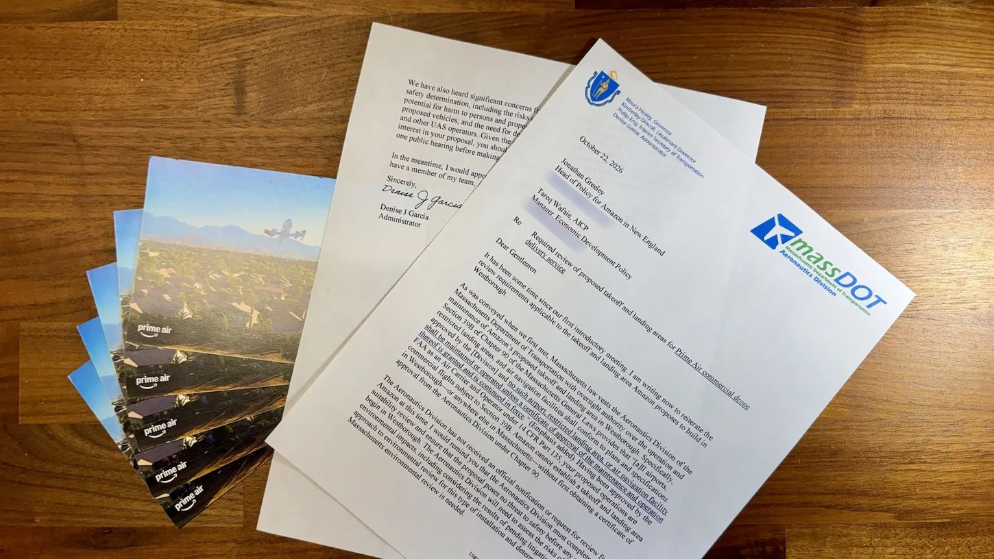 Printed two-page MassDOT Aeronautics Division letter dated October 22, 2026, on a wooden table next to a fanned stack of Amazon Prime Air brochures showing a delivery drone over houses