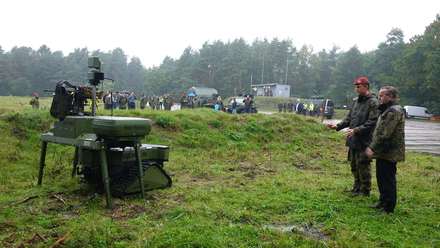 Boris Pistorius in a camouflage jacket stands beside a soldier in a red beret, looking at a green tracked ground robot fitted with a machine gun turret