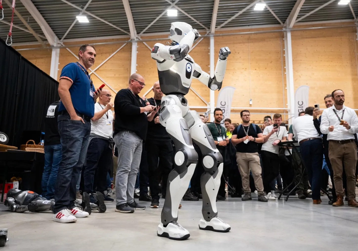 White humanoid robot raising one fist in a crowded exhibition hall while attendees film it on their phones