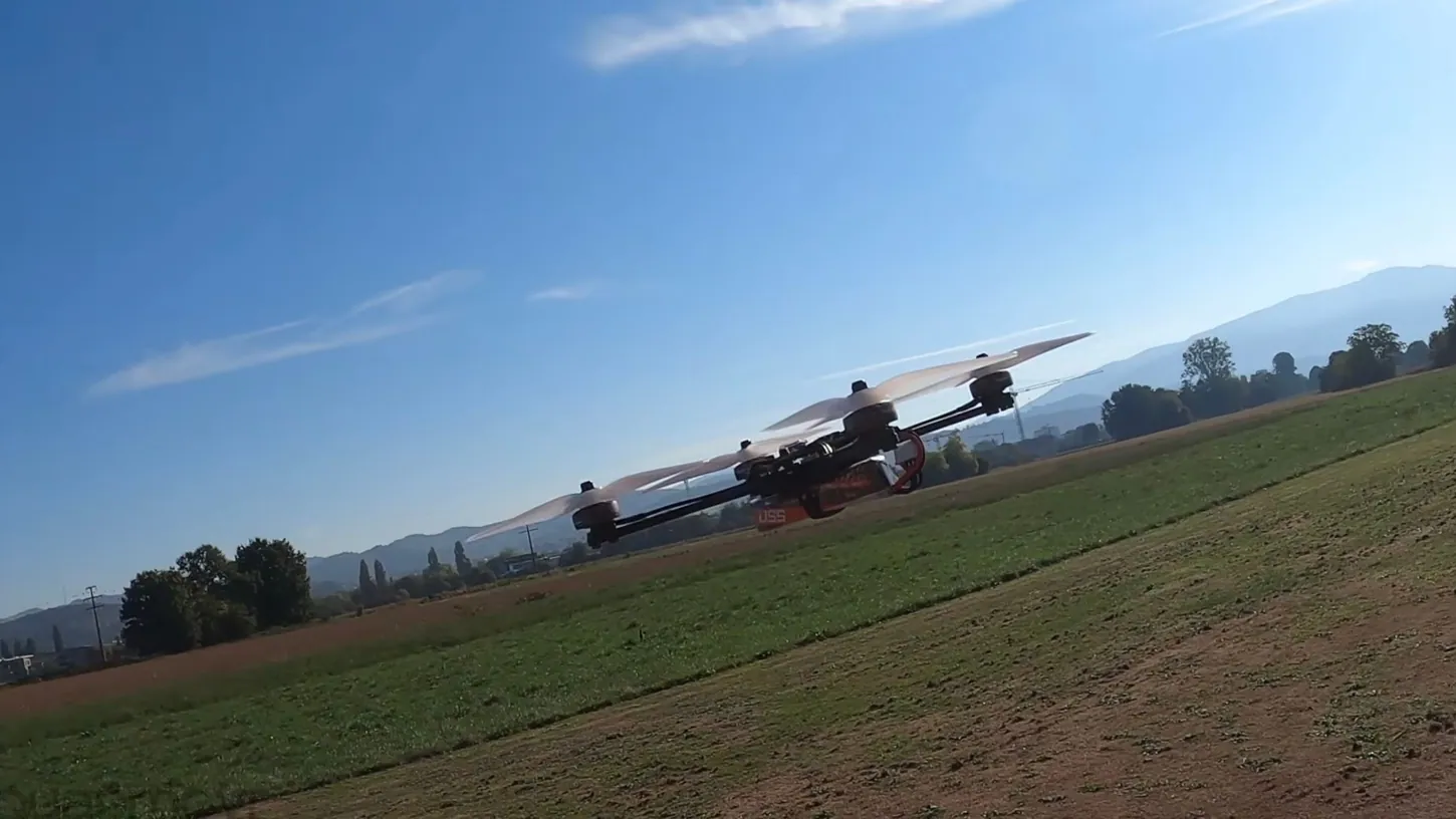 Small quadcopter with blurred propellers flying low over a mown field under a blue sky with hills behind