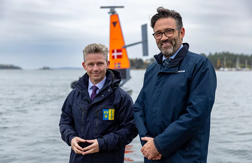 Swedish Defence Minister Pål Jonson and Saildrone's Robert Kleist, both in dark jackets, stand on a quay in front of an orange Saildrone sail