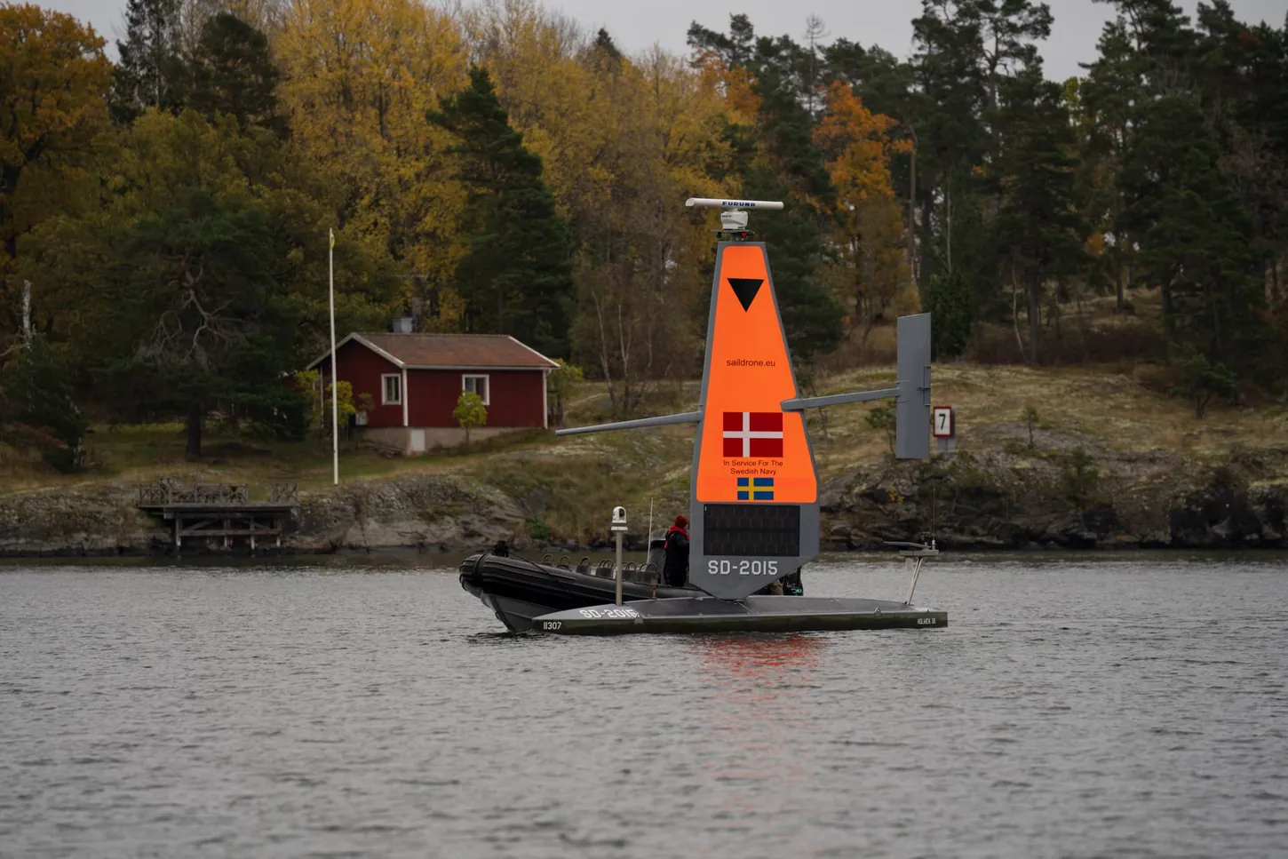 Saildrone Voyager SD-2015 with a black rigid inflatable boat alongside, in front of a red wooden cottage and autumn trees on a rocky shore