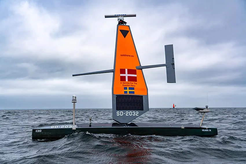 Saildrone Voyager SD-2032 under an orange sail marked In Service For The Swedish Navy, with Danish and Swedish flags, in choppy grey water