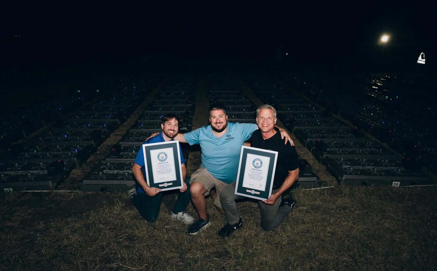 Three men kneeling on a dark field holding two framed Guinness World Records certificates in front of rows of drone storage trays