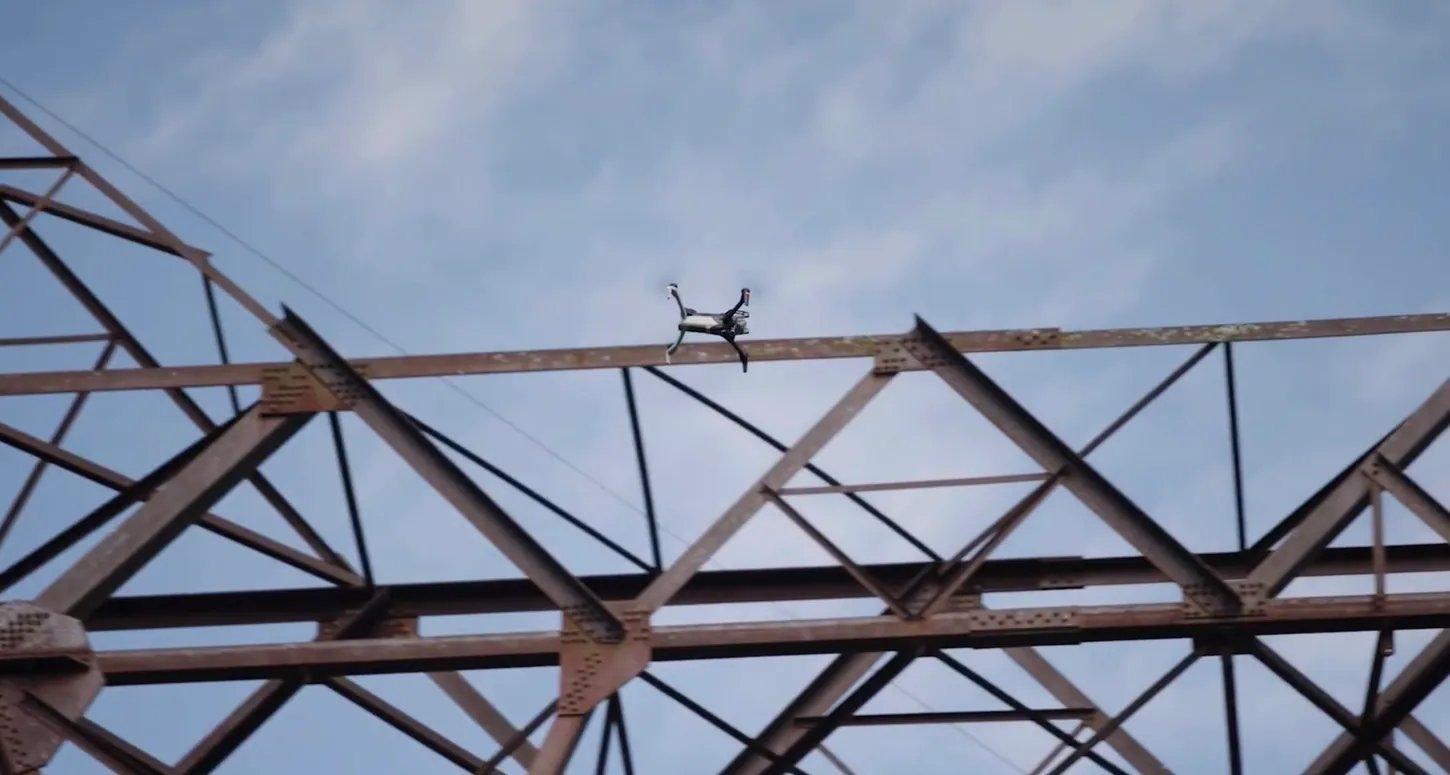 Skydio drone flying along the top beam of a rusted steel lattice transmission structure under a blue sky