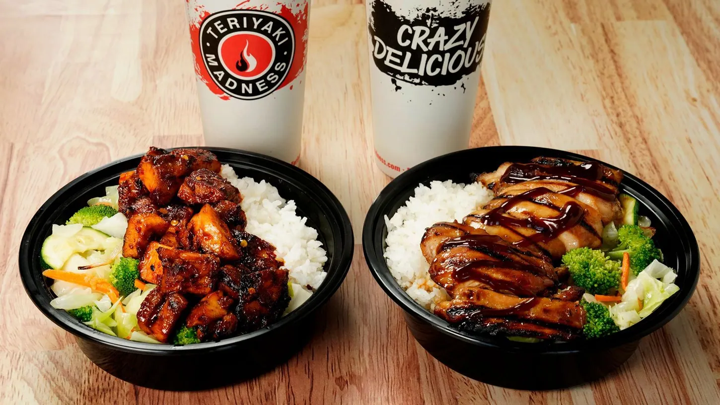 Two black bowls of grilled chicken teriyaki with rice, broccoli and cabbage on a wooden table, with two branded Teriyaki Madness drink cups behind them