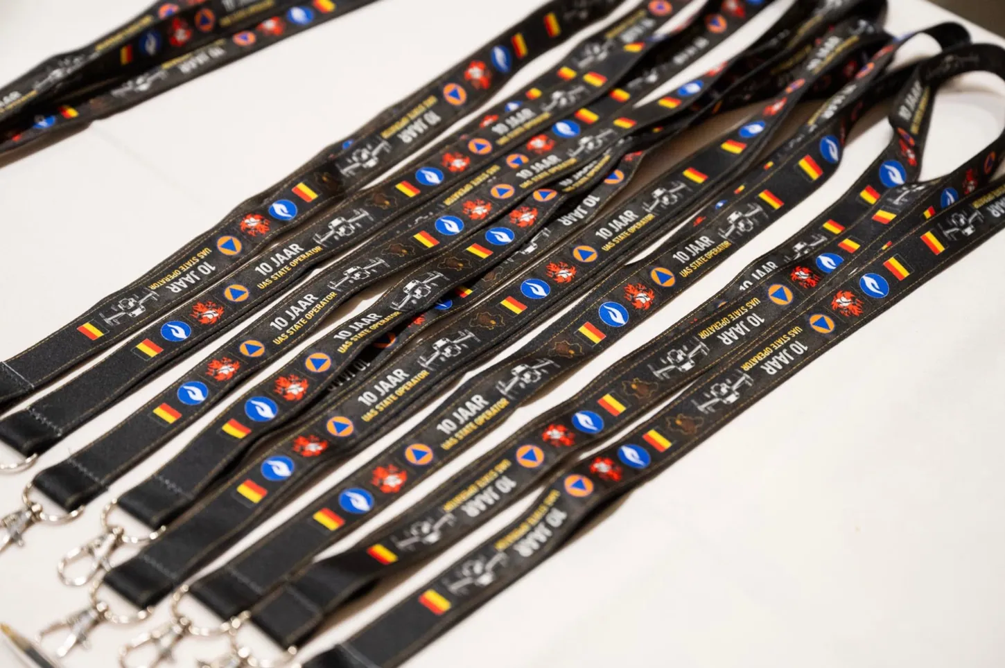 Black lanyards printed with 10 Jaar UAS State Operator, Belgian flags and police, fire and civil protection emblems laid out on a white table