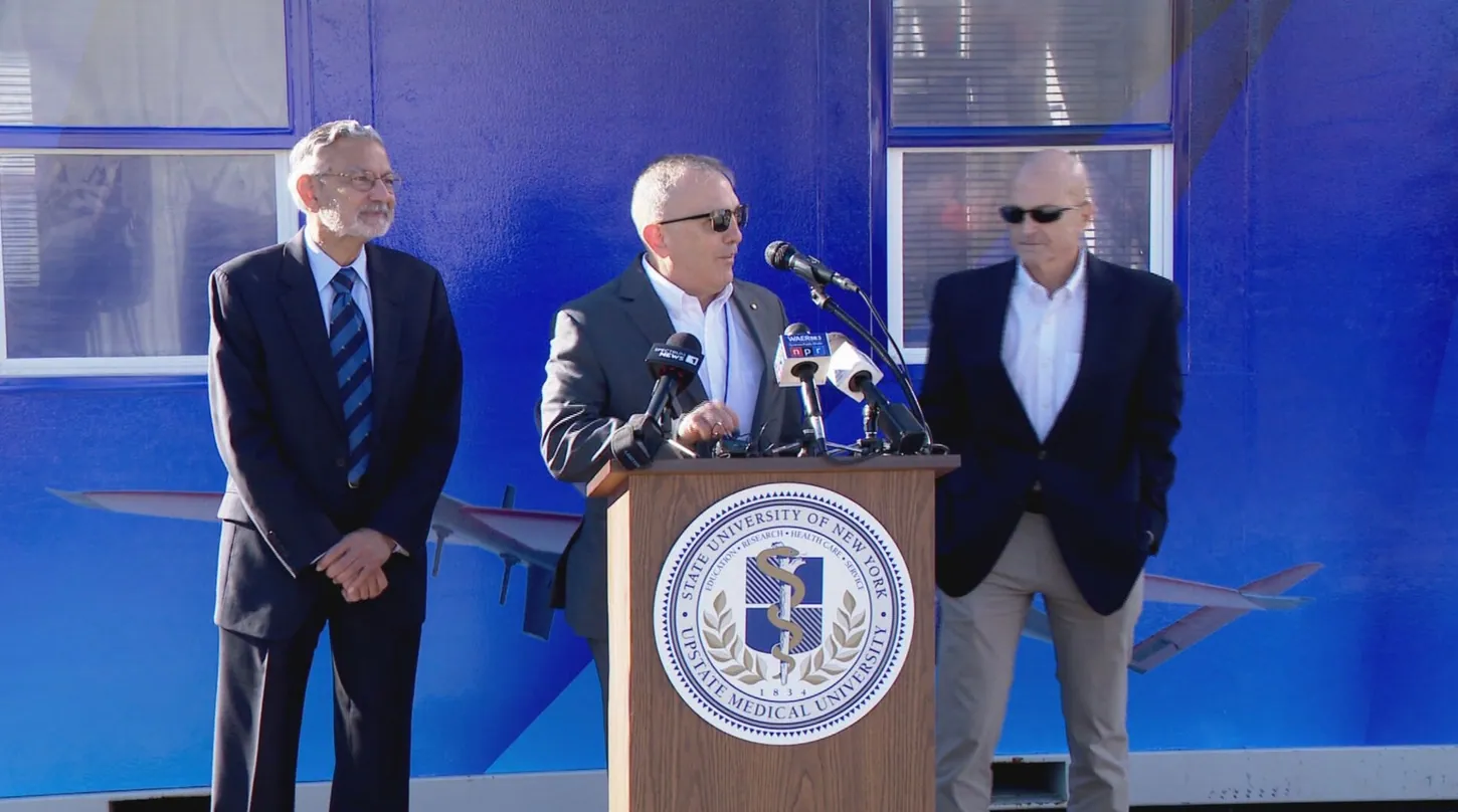 Three men at an outdoor press conference, one speaking at a podium bearing the State University of New York Upstate Medical University seal, in front of a blue building printed with drone graphics