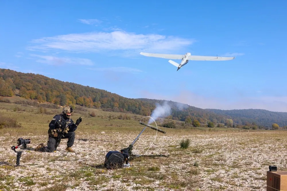 A Stork LR drone climbs away from a smoking pneumatic catapult on a gravel field while a kneeling US Army soldier watches it go