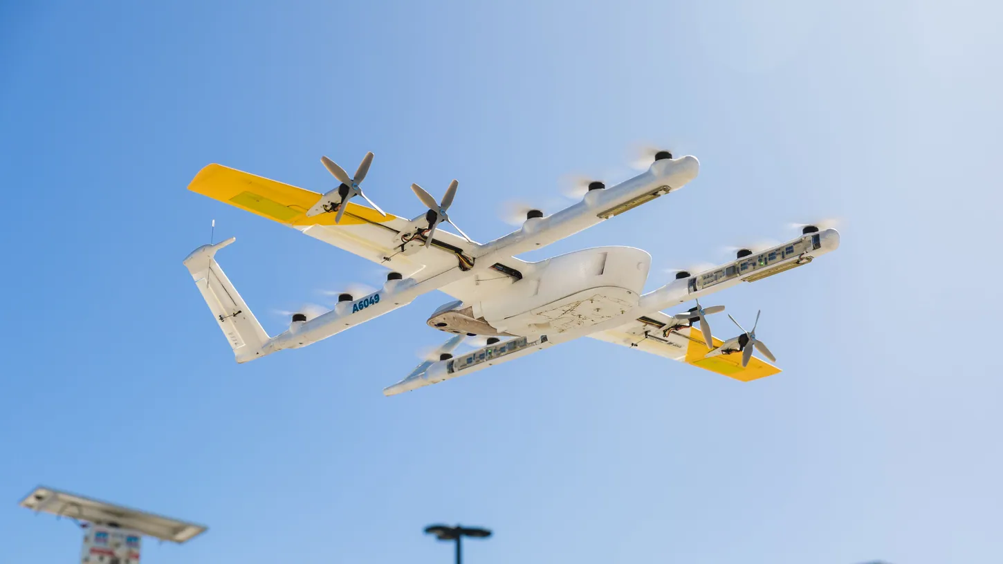 White and yellow Wing delivery drone hovering above a nest with landing pads and light poles below