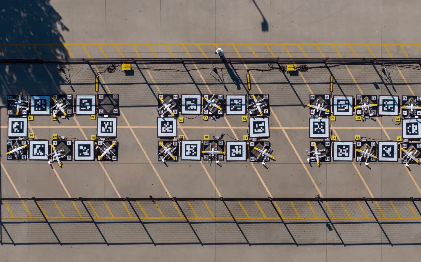 Aerial view of a Wing drone nest in a parking lot, with rows of charging pads and parked delivery drones inside a fence