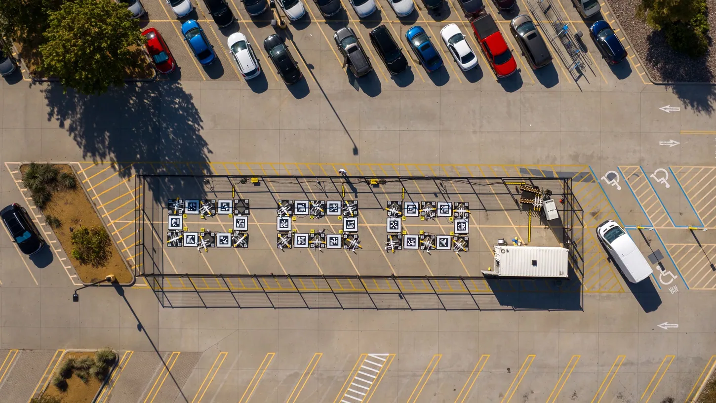 Aerial view of a fenced Wing drone nest with 18 landing pads and a white equipment container in a retail parking lot, surrounded by parked cars