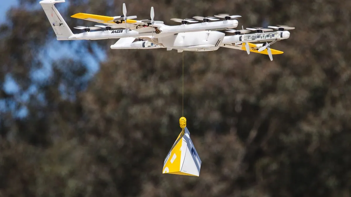 Wing delivery drone hovering above trees and lowering a yellow and white package on a tether