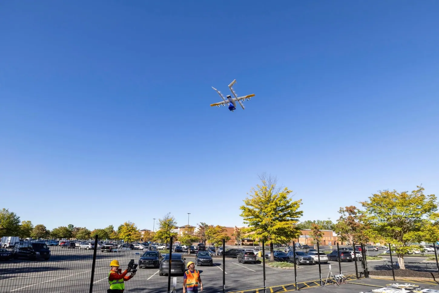 Wing delivery drone carrying a Walmart box flies over a fenced drone nest in a Walmart parking lot as two workers in hard hats and safety vests watch