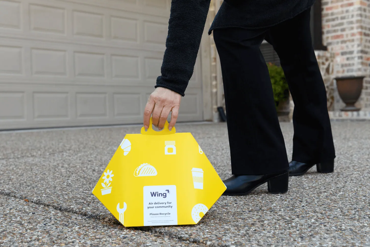 A person in black trousers and heeled boots bends to pick up a yellow Wing-branded delivery package from a driveway in front of a garage door