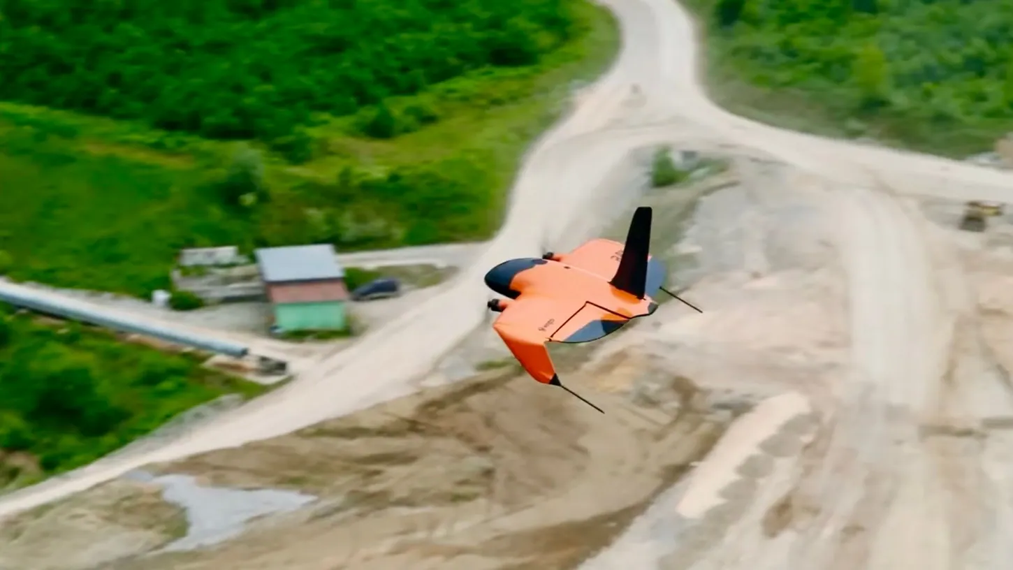 WingtraRAY banking over a dirt haul road between green hillsides, a small building and a pipeline below