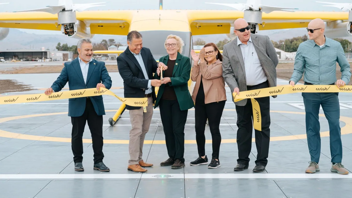 Six people holding a yellow Wisk ribbon on a vertiport landing pad, two of them cutting it with gold scissors, in front of a yellow Wisk air taxi