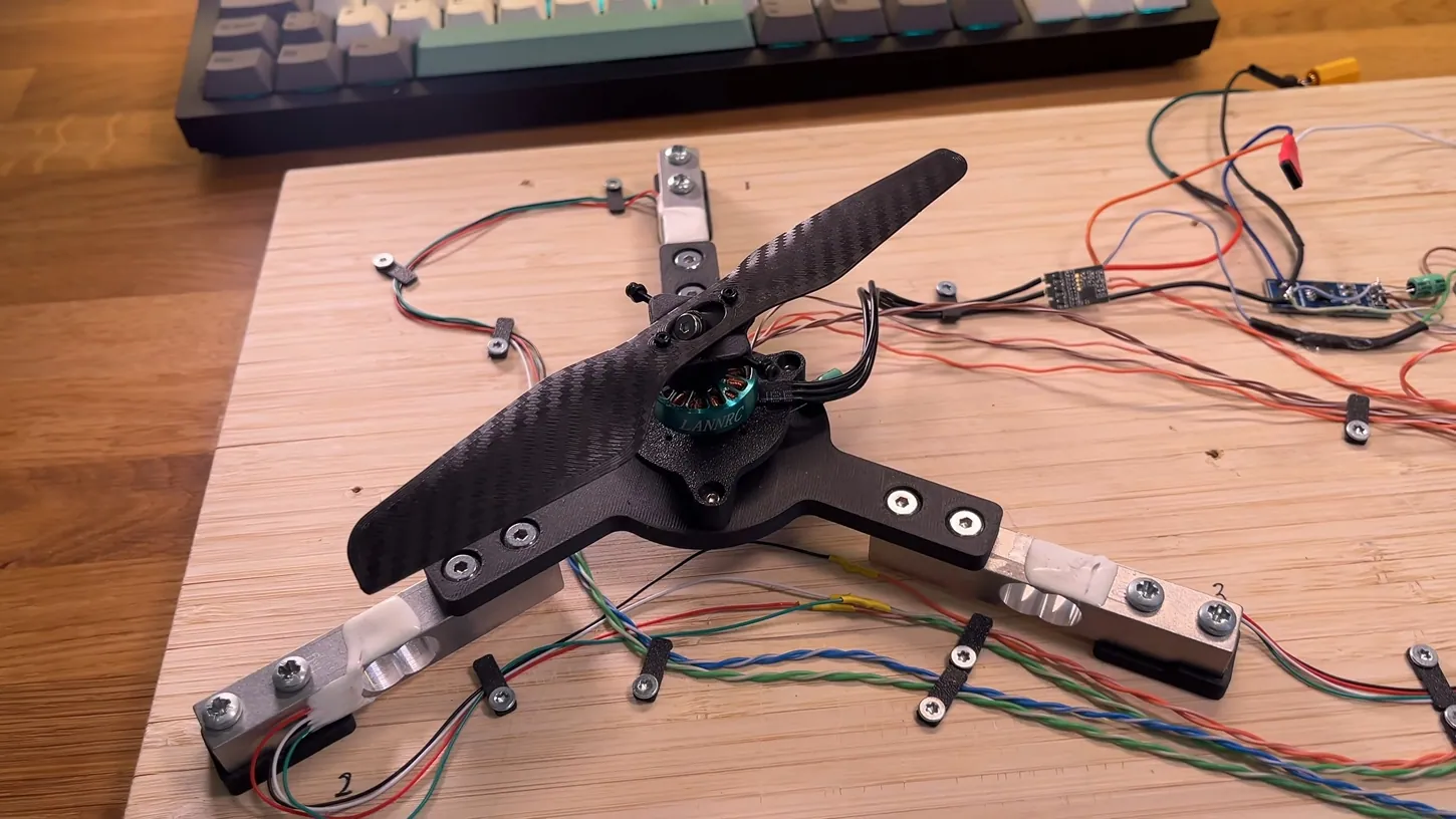 3D printed rotor with dark carbon-patterned blades on a black three-armed mount bolted to load cells on a wooden cutting board, with wiring running to a small controller