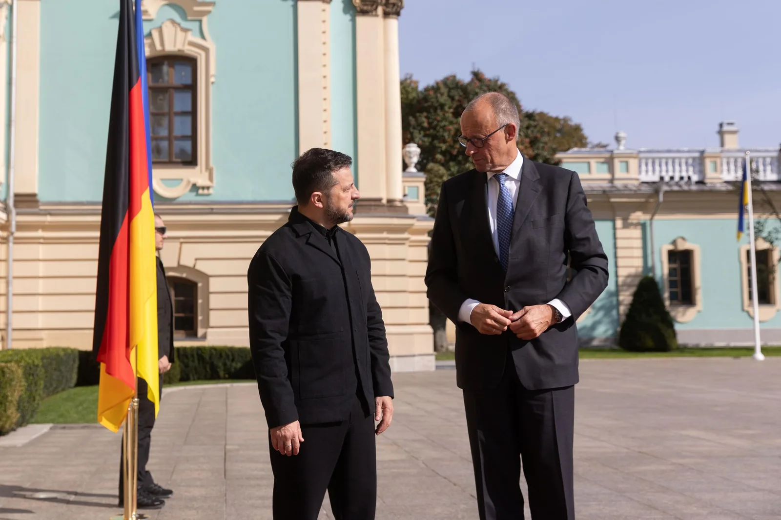Ukrainian President Volodymyr Zelensky and German Chancellor Friedrich Merz talking in front of the turquoise facade of the Mariinskyi Palace in Kyiv, a German flag beside them.
